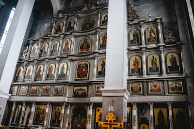 A large religious altar adorned with multiple framed icons depicting various saints and religious figures, set in a grand church interior. The icons are arranged in a structured, symmetric pattern on a multi-tiered wooden iconostasis. The walls are high and the structure has ornate decorative elements with intricate carvings.