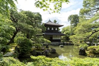 A serene Japanese garden featuring a traditional wooden pavilion surrounded by lush greenery and positioned next to a tranquil pond. The garden is adorned with carefully pruned trees and shrubs, and the water contains several rocks, enhancing the peaceful ambiance.
