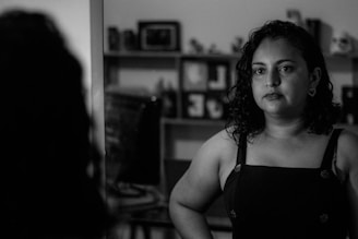 A person is looking at their reflection in a mirror. The scene is in black and white, creating a moody and introspective atmosphere. The background features a shelving unit with various objects, possibly books or decorative items.