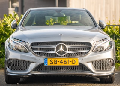 A sleek silver Mercedes-Benz car is parked, showcasing a shiny front grille with the iconic three-pointed star emblem. The vehicle features modern headlamps and a yellow license plate with European Union markings. Lush green foliage is visible in the background.