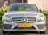 A sleek silver Mercedes-Benz car is parked, showcasing a shiny front grille with the iconic three-pointed star emblem. The vehicle features modern headlamps and a yellow license plate with European Union markings. Lush green foliage is visible in the background.