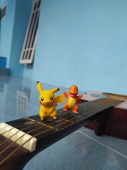 Two Pokémon figures, Pikachu and Charmander, are placed on the neck of a guitar. The background is a bright blue wall with a window.