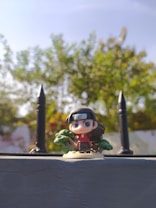A detailed anime-style figurine stands in the foreground against a blurred backdrop of lush green foliage under a bright blue sky. The figure features a headband with a symbol, long dark hair, and a red outfit. The scene is framed by spiked fence posts on either side.