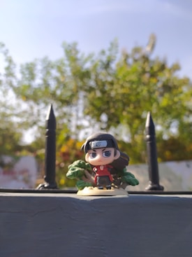 A detailed anime-style figurine stands in the foreground against a blurred backdrop of lush green foliage under a bright blue sky. The figure features a headband with a symbol, long dark hair, and a red outfit. The scene is framed by spiked fence posts on either side.