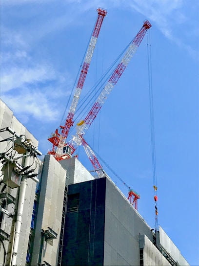 Two large cranes with red and white structures rise above a high-rise building under construction. The building is partially covered, with electrical cables and installations visible on its exterior. The sky is bright and clear.
