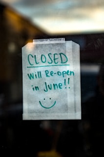 A handwritten note taped to a glass door or window, stating 'CLOSED Will Re-open in June!!' with a smiley face underneath. The sign is written with green marker on a white piece of paper and is taped at the top corners.