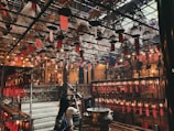 An intricately designed interior with numerous hanging spiral incense coils adorned with red paper tags. The space is dimly lit with warm yellow lights, casting a cozy and calm atmosphere. A woman is observing the surroundings, which include traditional wooden structures and ornate details, suggesting a spiritual or cultural setting.