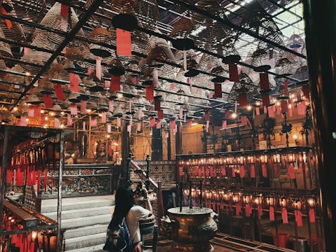 An intricately designed interior with numerous hanging spiral incense coils adorned with red paper tags. The space is dimly lit with warm yellow lights, casting a cozy and calm atmosphere. A woman is observing the surroundings, which include traditional wooden structures and ornate details, suggesting a spiritual or cultural setting.