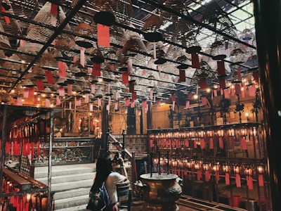 An intricately designed interior with numerous hanging spiral incense coils adorned with red paper tags. The space is dimly lit with warm yellow lights, casting a cozy and calm atmosphere. A woman is observing the surroundings, which include traditional wooden structures and ornate details, suggesting a spiritual or cultural setting.