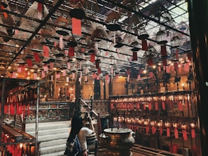 An intricately designed interior with numerous hanging spiral incense coils adorned with red paper tags. The space is dimly lit with warm yellow lights, casting a cozy and calm atmosphere. A woman is observing the surroundings, which include traditional wooden structures and ornate details, suggesting a spiritual or cultural setting.