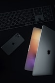 A minimalist arrangement featuring a laptop partially open, a smartphone, and a keyboard, all showing a sleek and modern design. The laptop's screen emits a soft glow of multicolored lights.