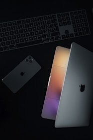 A minimalist arrangement featuring a laptop partially open, a smartphone, and a keyboard, all showing a sleek and modern design. The laptop's screen emits a soft glow of multicolored lights.