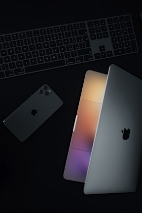 A minimalist arrangement featuring a laptop partially open, a smartphone, and a keyboard, all showing a sleek and modern design. The laptop's screen emits a soft glow of multicolored lights.