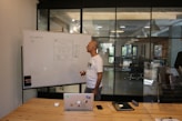 A man stands at a whiteboard in a modern office setting. The whiteboard displays a website wireframe sketch with some notes. The man is wearing a white t-shirt and is holding a marker. In front of him, on a wooden table, there is a laptop with various stickers, a smartphone, and an electronic device. The office features large windows and sleek lighting fixtures.