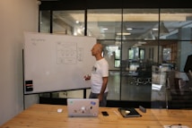 A man stands at a whiteboard in a modern office setting. The whiteboard displays a website wireframe sketch with some notes. The man is wearing a white t-shirt and is holding a marker. In front of him, on a wooden table, there is a laptop with various stickers, a smartphone, and an electronic device. The office features large windows and sleek lighting fixtures.