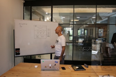 A man stands at a whiteboard in a modern office setting. The whiteboard displays a website wireframe sketch with some notes. The man is wearing a white t-shirt and is holding a marker. In front of him, on a wooden table, there is a laptop with various stickers, a smartphone, and an electronic device. The office features large windows and sleek lighting fixtures.