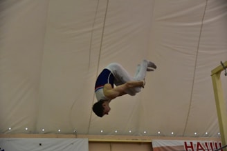 A gymnast performs a skill, mid-air, with a focus on acrobatics under a large indoor tent. The gymnast wears a competition uniform and is captured in a dynamic pose, showcasing control and strength.