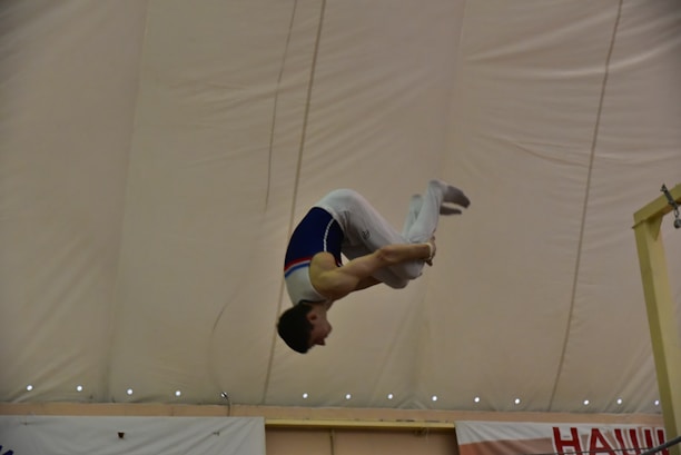 A gymnast performs a skill, mid-air, with a focus on acrobatics under a large indoor tent. The gymnast wears a competition uniform and is captured in a dynamic pose, showcasing control and strength.