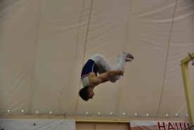 A gymnast performs a skill, mid-air, with a focus on acrobatics under a large indoor tent. The gymnast wears a competition uniform and is captured in a dynamic pose, showcasing control and strength.