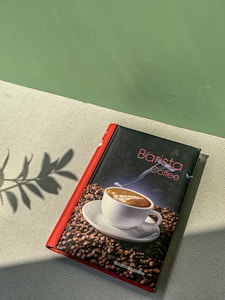 A book with a red spine titled 'Barista Coffee' rests on a light-textured surface. Its cover features an image of a steaming cup of latte art surrounded by coffee beans. A shadow of leaves is cast on the surface beside the book, with a green backdrop adding contrast.
