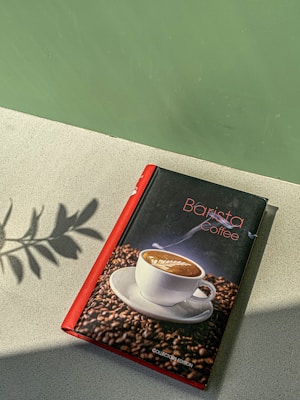 A book with a red spine titled 'Barista Coffee' rests on a light-textured surface. Its cover features an image of a steaming cup of latte art surrounded by coffee beans. A shadow of leaves is cast on the surface beside the book, with a green backdrop adding contrast.