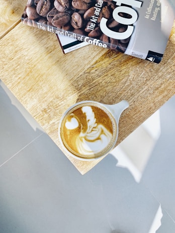 A cappuccino in a white cup displays intricate latte art on a light wooden table, next to a coffee-themed book titled 'The Art and Craft of Coffee', which features an image of coffee beans on its cover.