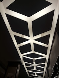 A geometric ceiling design featuring a series of interconnected rectangular light panels arranged in a repetitive pattern. The lights create a contrasting effect against the dark ceiling background.