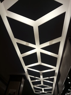 A geometric ceiling design featuring a series of interconnected rectangular light panels arranged in a repetitive pattern. The lights create a contrasting effect against the dark ceiling background.
