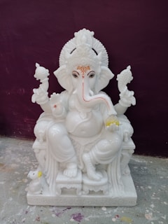 A white marble statue of a deity with the head of an elephant, characterized by intricate detailing on the headdress and jewelry. The figure is seated on a throne with one leg bent and the other resting on the base. It holds objects in its hands, symbolizing religious or cultural significance. A small mouse figure is positioned beside the statue on the ground.