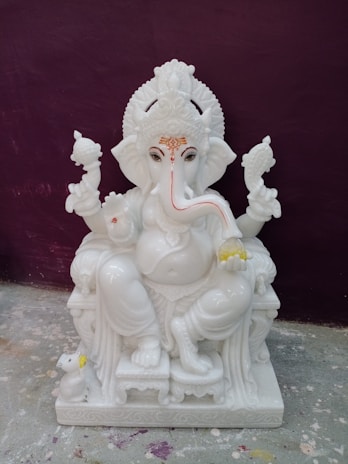 A white marble statue of a deity with the head of an elephant, characterized by intricate detailing on the headdress and jewelry. The figure is seated on a throne with one leg bent and the other resting on the base. It holds objects in its hands, symbolizing religious or cultural significance. A small mouse figure is positioned beside the statue on the ground.