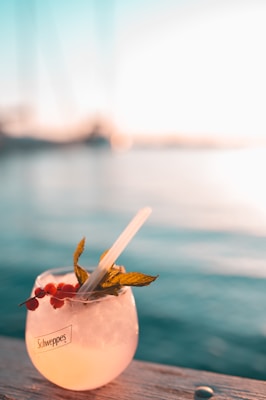 A refreshing beverage in a glass sits on a wooden surface with a backdrop of a body of water. The drink is garnished with small red berries, fresh mint leaves, and has a clear straw. The image conveys a warm, inviting atmosphere, enhanced by the soft, golden sunlight reflecting off the surface of the water.