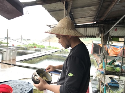 A man in a traditional straw hat is standing near a fish farm, holding a metal pot with food. He is surrounded by water tanks and various fishing equipment. The setting appears to be rural, with makeshift structures providing some shelter.