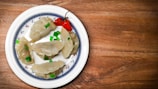 A plate of dumplings with chopped green onions and a red chili pepper arranged on a decorative white dish. The dumplings have a translucent, soft appearance and sit on a wooden table.