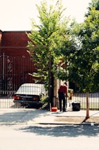 A person wearing a red shirt is standing near a gate, holding a broom and cleaning the sidewalk. A black sedan is parked behind the gate, next to a brick building. Trees with green leaves provide shade, and a small orange object, possibly a toolbox, is on the ground nearby.