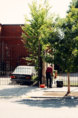 A person wearing a red shirt is standing near a gate, holding a broom and cleaning the sidewalk. A black sedan is parked behind the gate, next to a brick building. Trees with green leaves provide shade, and a small orange object, possibly a toolbox, is on the ground nearby.
