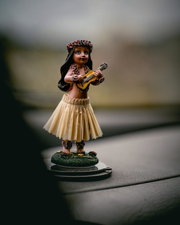 A small hula dancer figurine with long hair, wearing a traditional grass skirt and floral accessories, holds a small ukulele. The scene is captured on a car dashboard with a blurred background.