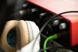 Close-up view of audio equipment featuring a red audio interface and beige headphones. Cables are connected to the interface, and various knobs are visible. The lighting is soft, creating a warm atmosphere.