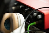 Close-up view of audio equipment featuring a red audio interface and beige headphones. Cables are connected to the interface, and various knobs are visible. The lighting is soft, creating a warm atmosphere.