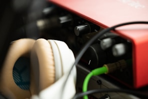 Close-up view of audio equipment featuring a red audio interface and beige headphones. Cables are connected to the interface, and various knobs are visible. The lighting is soft, creating a warm atmosphere.