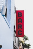 A vertical red banner is mounted on the exterior of a building, displaying the letters BARC in black. The building is painted with light blue bricks, and there is a cylindrical speaker or light fixture attached to it. Part of the city street and a tree are visible in the background.