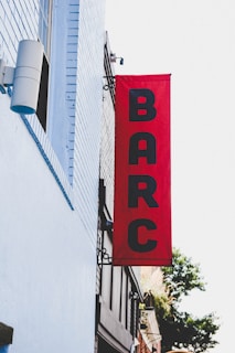 A vertical red banner is mounted on the exterior of a building, displaying the letters BARC in black. The building is painted with light blue bricks, and there is a cylindrical speaker or light fixture attached to it. Part of the city street and a tree are visible in the background.
