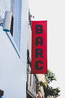 A vertical red banner is mounted on the exterior of a building, displaying the letters BARC in black. The building is painted with light blue bricks, and there is a cylindrical speaker or light fixture attached to it. Part of the city street and a tree are visible in the background.