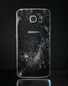 A smartphone with a severely cracked back panel, revealing a spiderweb-like pattern of cracks. The camera and flash are intact, but the damage is extensive.