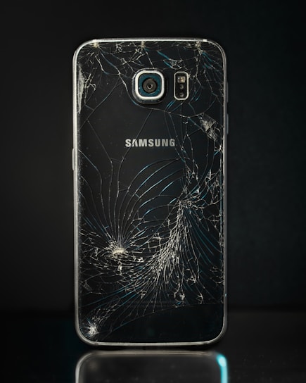 A smartphone with a severely cracked back panel, revealing a spiderweb-like pattern of cracks. The camera and flash are intact, but the damage is extensive.