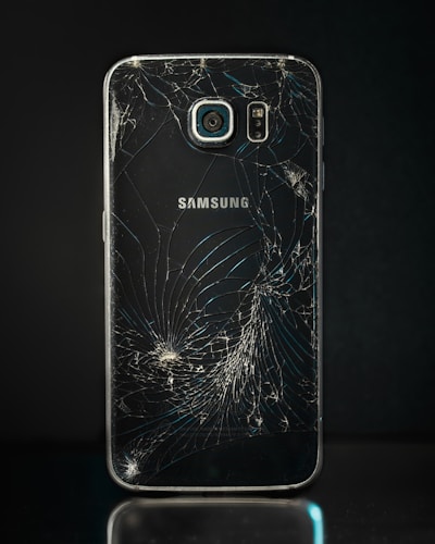 A smartphone with a severely cracked back panel, revealing a spiderweb-like pattern of cracks. The camera and flash are intact, but the damage is extensive.