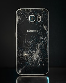 A smartphone with a severely cracked back panel, revealing a spiderweb-like pattern of cracks. The camera and flash are intact, but the damage is extensive.