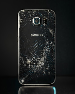 A smartphone with a severely cracked back panel, revealing a spiderweb-like pattern of cracks. The camera and flash are intact, but the damage is extensive.