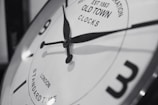 A close-up of a vintage-style clock face with black hour and minute hands pointing towards numbers. The clock has a reflective metallic rim and features text indicating 'Old Town Clocks' with 'London Standard Time'. The overall image is in black and white, highlighting the classic design.