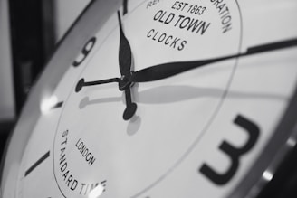A close-up of a vintage-style clock face with black hour and minute hands pointing towards numbers. The clock has a reflective metallic rim and features text indicating 'Old Town Clocks' with 'London Standard Time'. The overall image is in black and white, highlighting the classic design.
