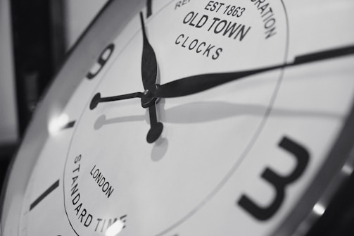 A close-up of a vintage-style clock face with black hour and minute hands pointing towards numbers. The clock has a reflective metallic rim and features text indicating 'Old Town Clocks' with 'London Standard Time'. The overall image is in black and white, highlighting the classic design.